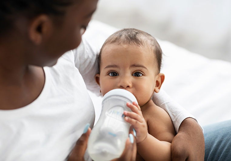 Nursery And Feeding Concept. Closeup of adorable small black baby drinking from bottle, young smiling woman holding kid on hands, taking care of her child, sitting on bed at home, selective focus