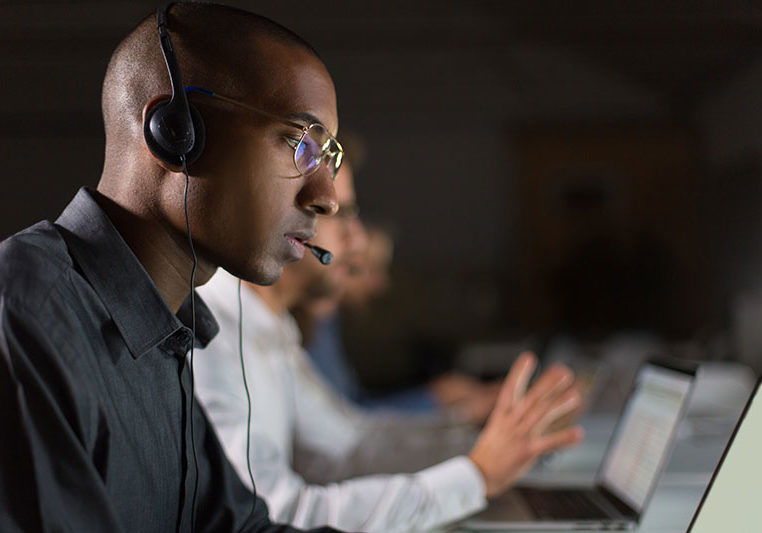 Focused call center operator communicating with client. African American young man in eyeglasses looking at laptop while serving client. Call center concept