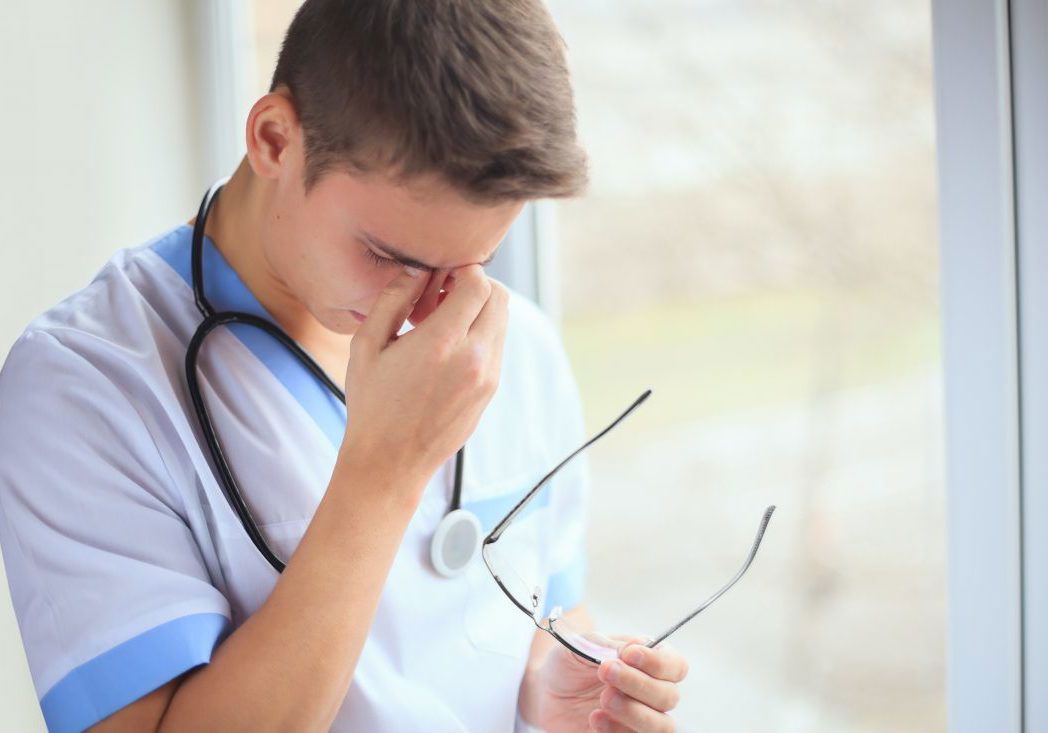 Portrait of unhappy tired young doctor having headache near a window